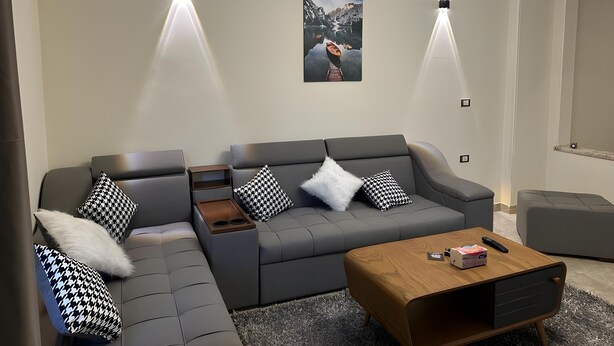 A modern living room with a grey sectional sofa, black and white patterned pillows, a wooden coffee table, and a mounted artwork of a mountainous landscape. - City Center: Modern 2BR Flat-56 (Cairo Governorate)