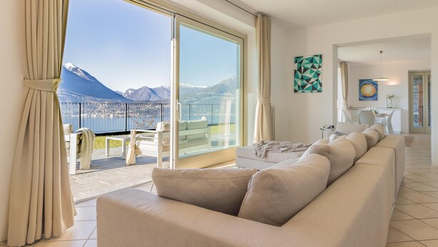 A modern living room with a large beige sofa, a dining area with a table and chairs, and a balcony with a view of mountains and a lake. - Varenna O Sole Mio in Perledo (Perledo)