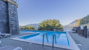 A rectangular pool with two ladders, surrounded by lounge chairs, a stone wall, and a view of mountains. - Varenna O Sole Mio in Perledo (Perledo)