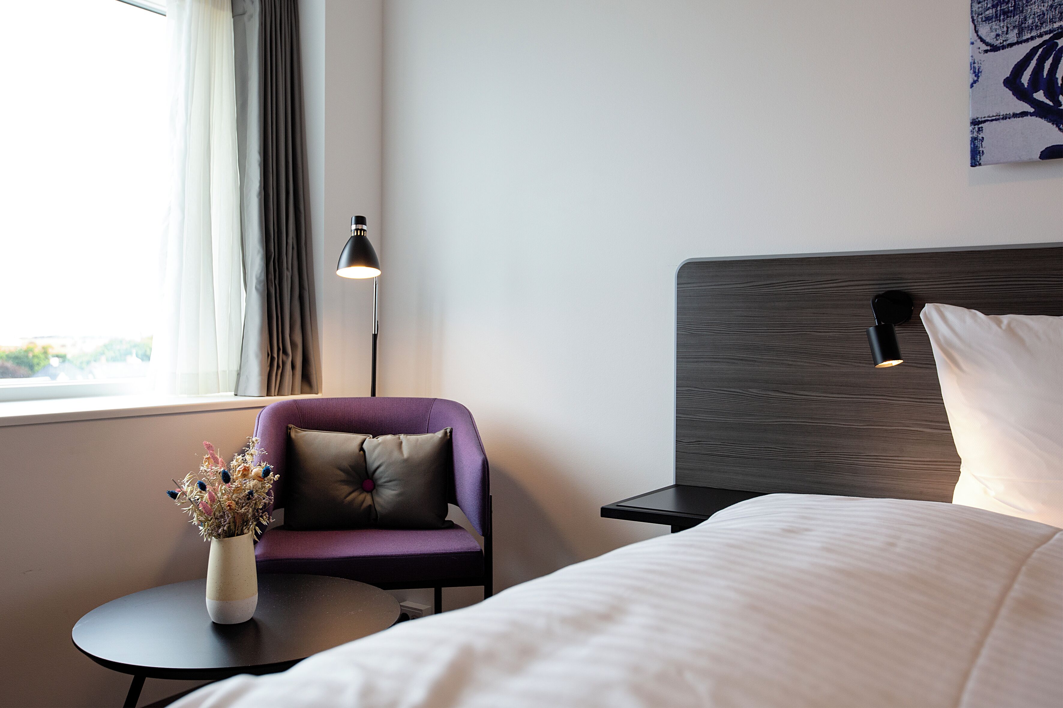 A hotel room with a bed, a small table with a vase of flowers, a purple armchair, a lamp, and a painting on the wall.