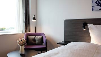 A hotel room with a bed, a small table with a vase of flowers, a purple armchair, a lamp, and a painting on the wall.