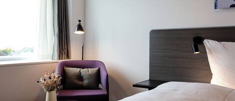 A hotel room with a bed, a small table with a vase of flowers, a purple armchair, a lamp, and a painting on the wall.