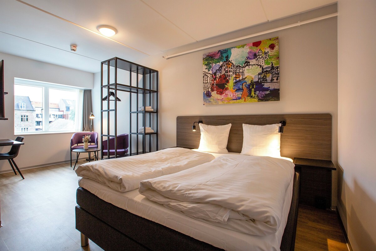 A modern hotel room with a large bed, a desk, a chair, and a window with a view of buildings.