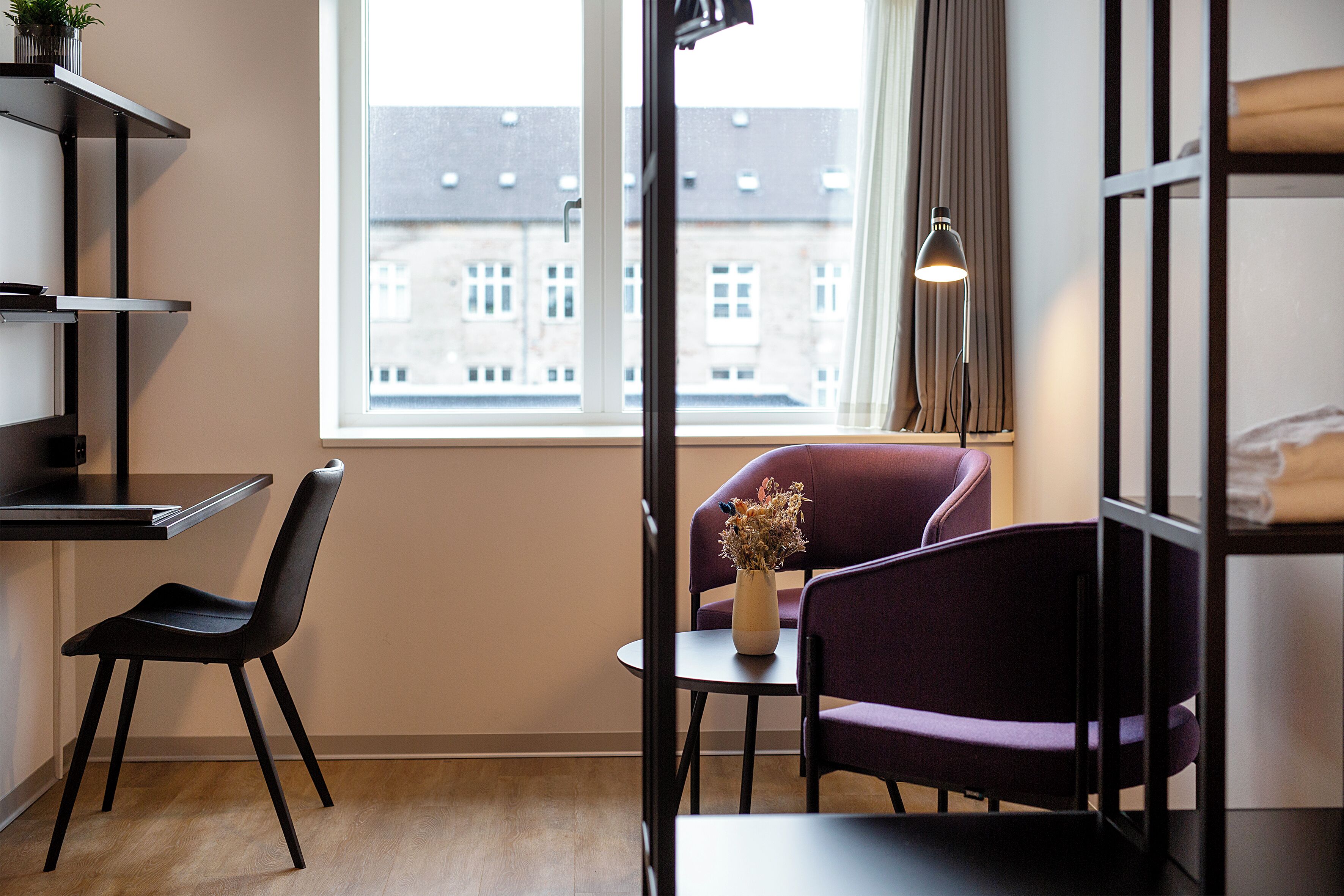 A modern room with a wooden floor, a black chair, a purple chair, a small table with a vase of dried flowers, and a window with a view of buildings.