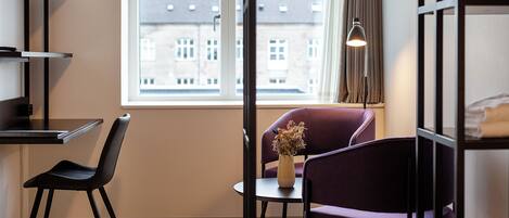 A modern room with a wooden floor, a black chair, a purple chair, a small table with a vase of dried flowers, and a window with a view of buildings.