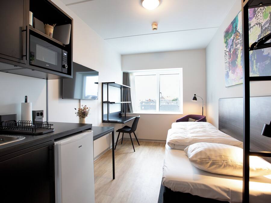 Habitación de hotel compacta con kitchenette, cama individual, escritorio y ventana que proporciona luz natural.