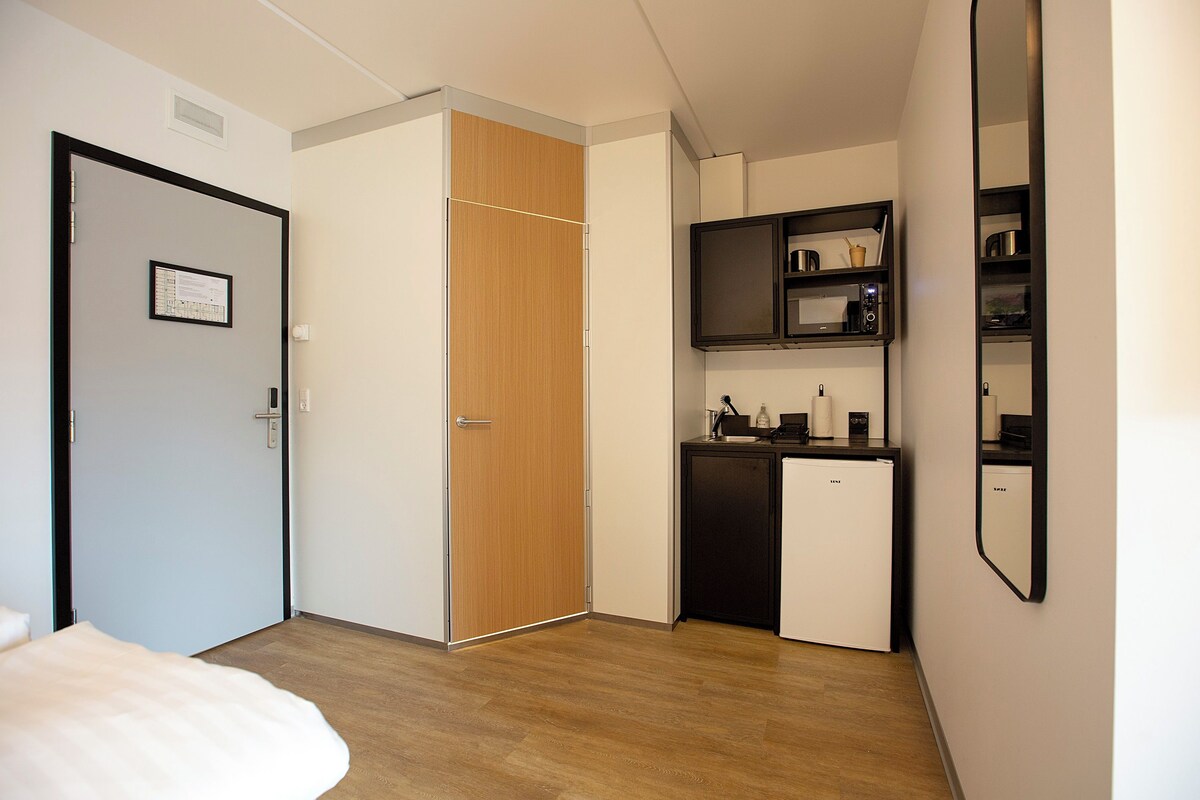 Ein gemütlicher Raum mit Bett, Holzschrank, einer kleinen Küchenzeile mit Mikrowelle und einer integrierten Regaleinheit.