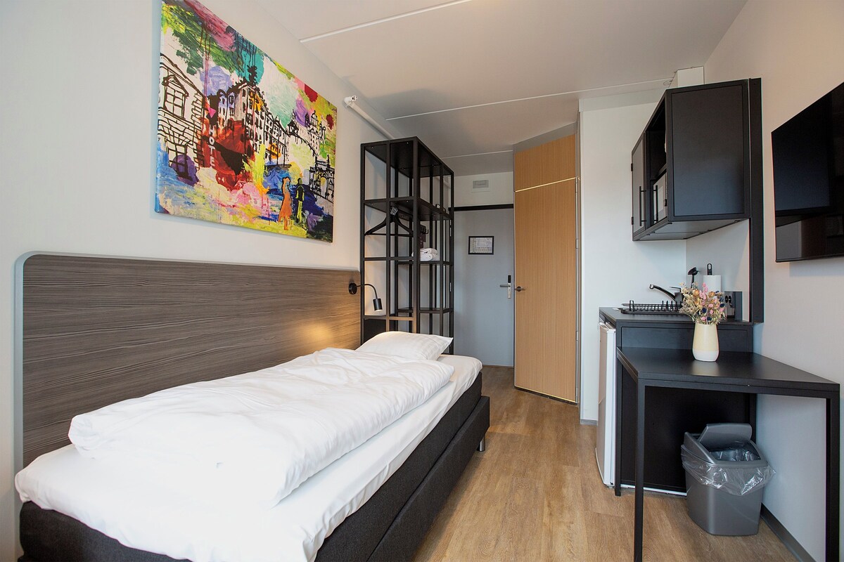 A modern hotel room with a bed, a desk, a trash bin, and a colorful painting on the wall.