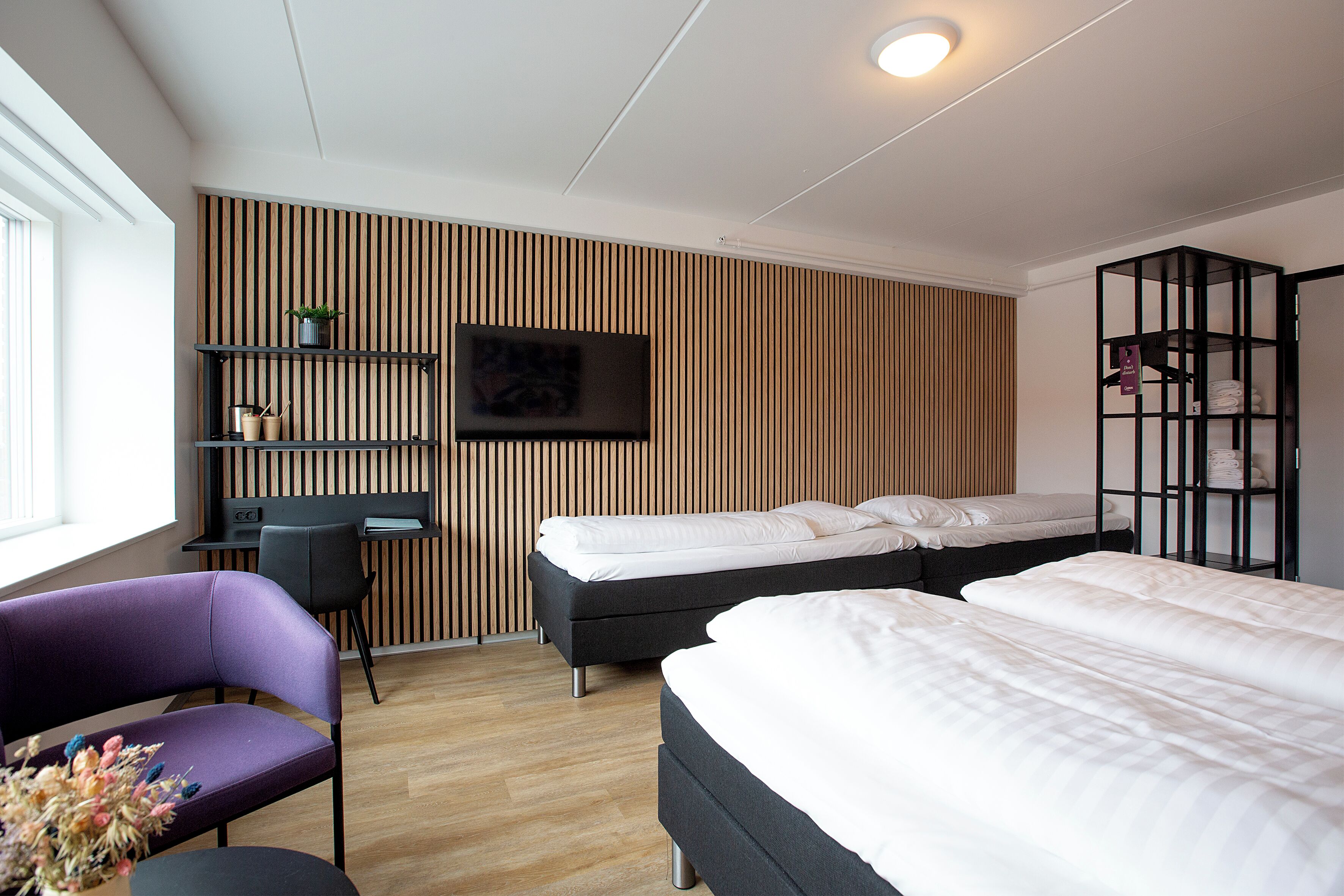 A modern hotel room with a wooden slat wall, a TV mounted above a desk, a purple chair, two beds, and a black shelving unit.