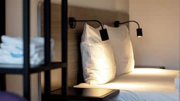 Cama bem arrumada com lençóis brancos e duas almofadas, mesa de cabeceira com abajur e cabeceira de madeira.