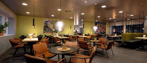 A modern restaurant interior with round tables and brown chairs, featuring overhead lighting and a vibrant mural on the wall.