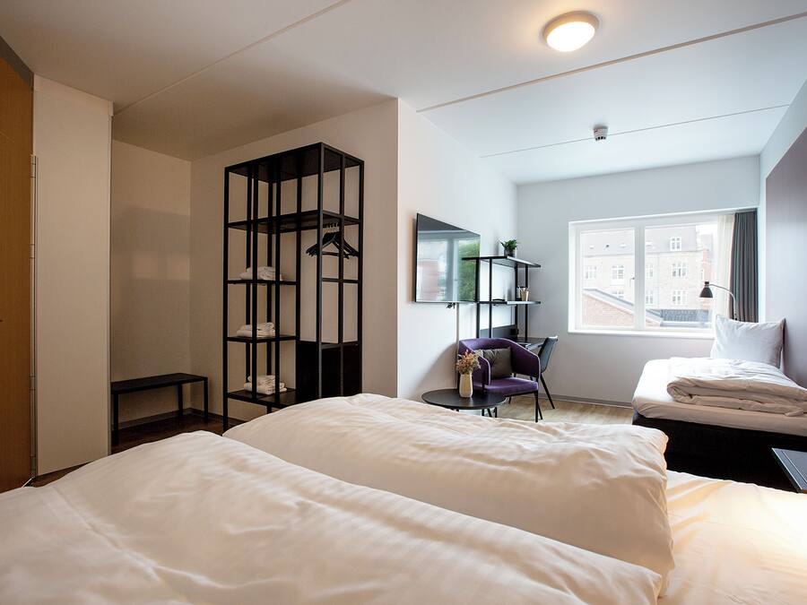 A hotel room with two beds, a desk, a chair, a wardrobe, and a window with a view of buildings.
