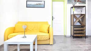 Un sofá amarillo brillante con una mesa blanca al frente, un estante de madera con vasos, un cuadro enmarcado en la pared y una puerta con marco verde.