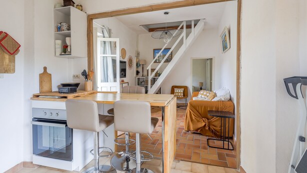 Une cuisine avec une table en bois et des tabourets de bar, un four blanc et un escalier menant à un espace de vie comprenant un canapé et un lit.