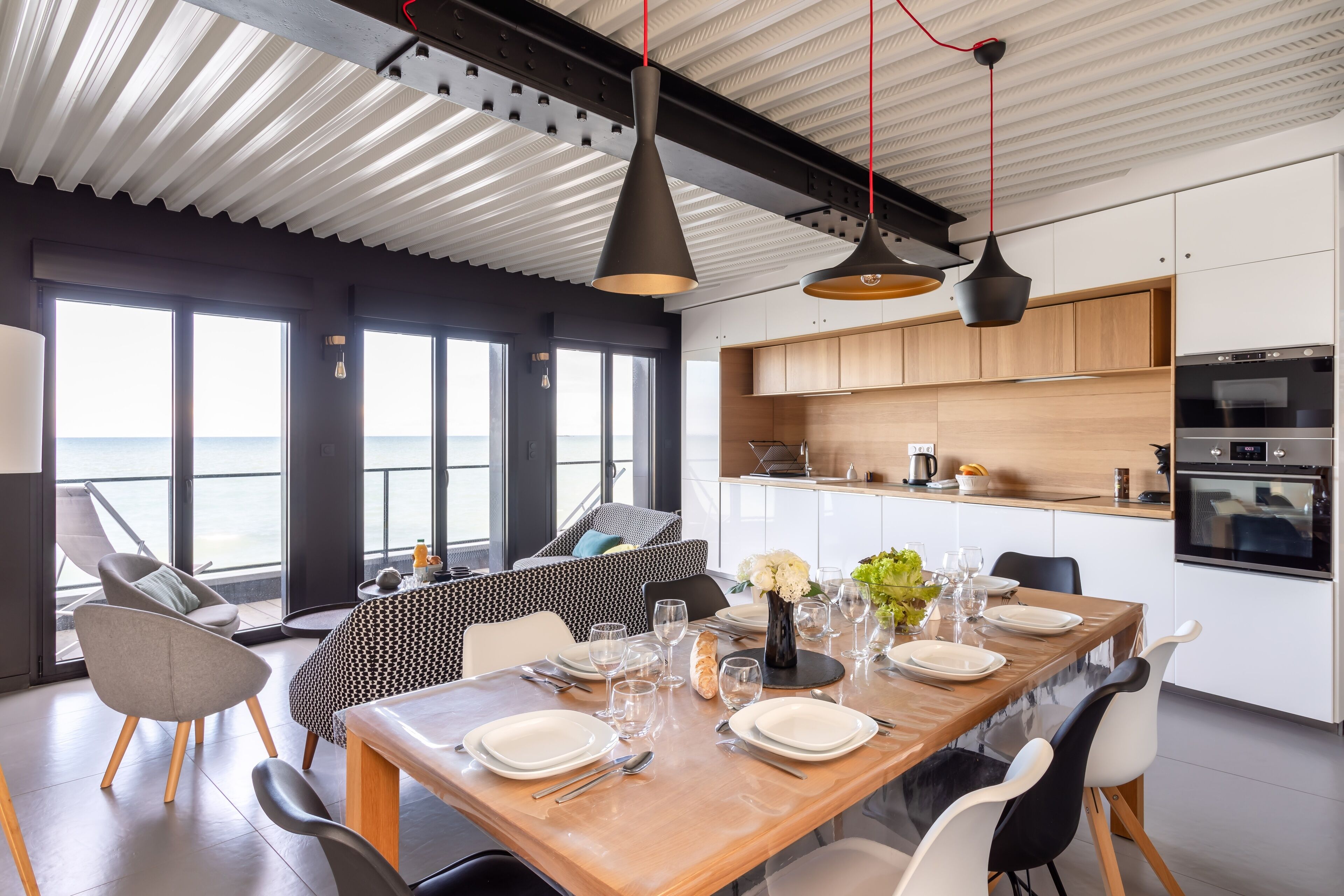 Un espace repas moderne avec une table en bois pouvant accueillir six personnes, de grandes fenêtres offrant une vue sur la mer, et une cuisine dotée d’armoires en bois et d’appareils électroménagers en acier inoxydable.