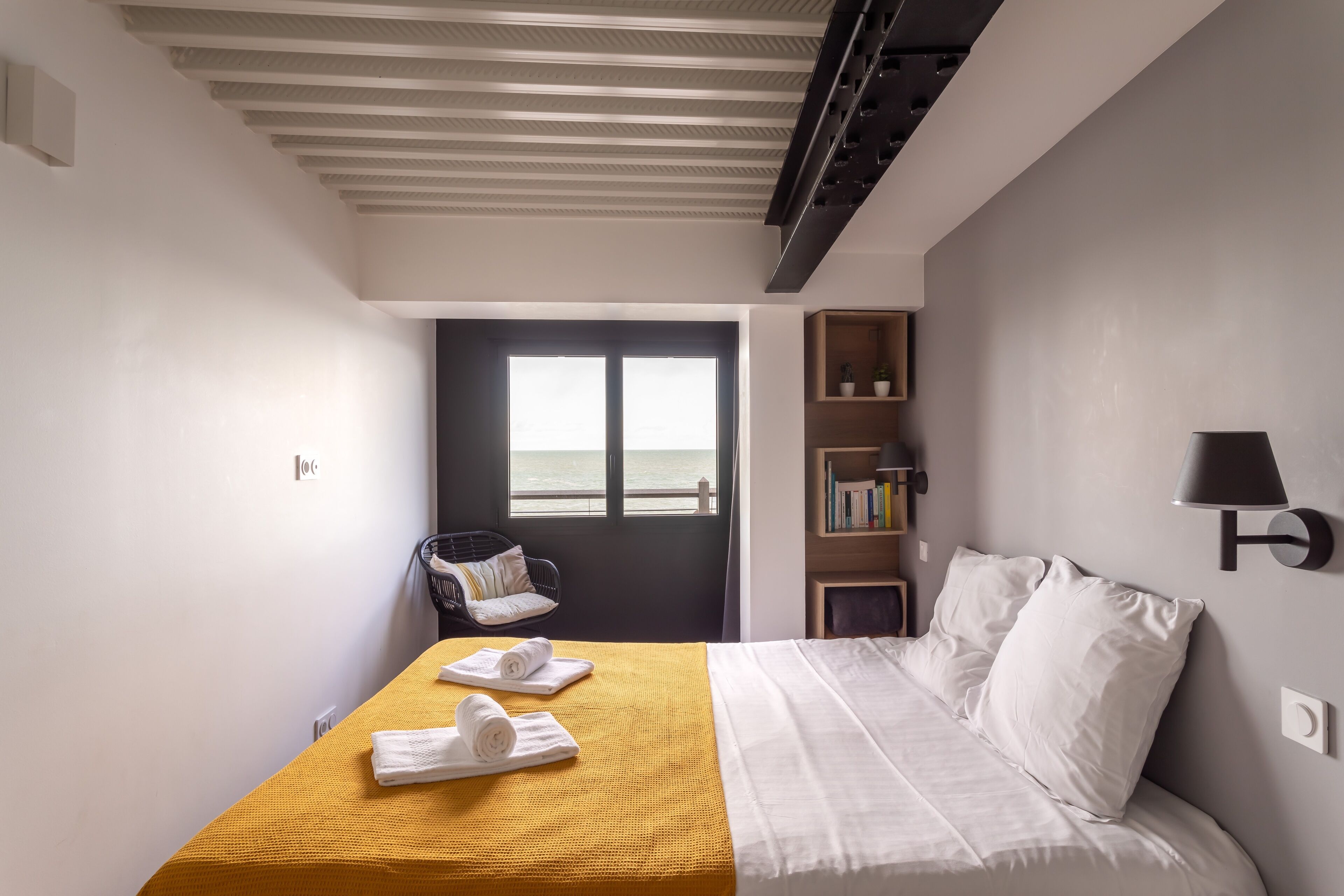 Une chambre d’hôtel avec un lit, une couvre-lit jaune, une chaise, une bibliothèque et une vue sur la mer par la fenêtre.