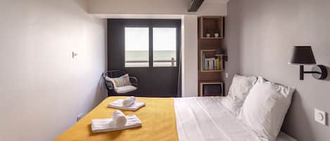 Une chambre d’hôtel avec un lit, une couvre-lit jaune, une chaise, une bibliothèque et une vue sur la mer par la fenêtre.