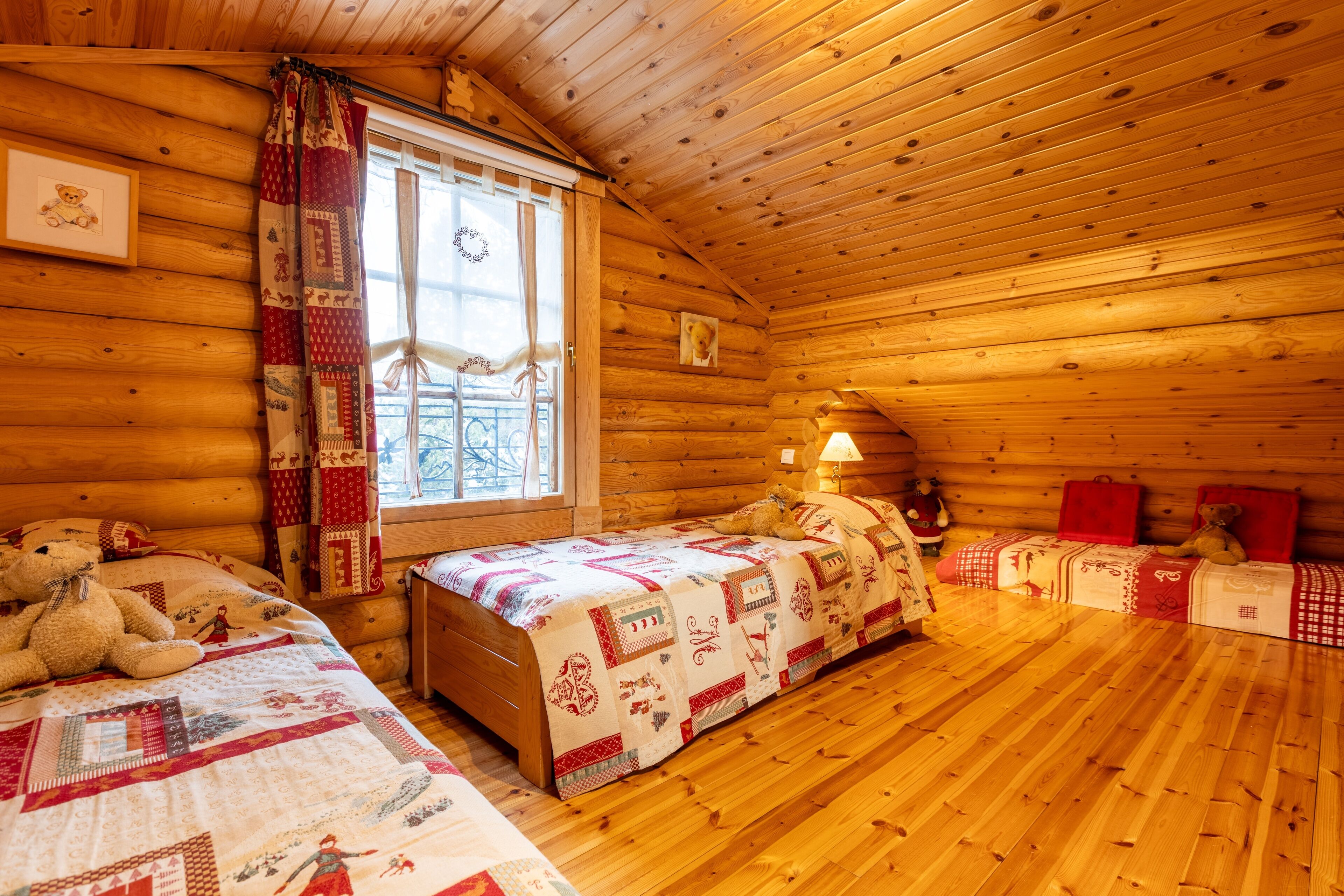 A cozy log cabin bedroom with two beds, a window with curtains, and a teddy bear on one bed.
