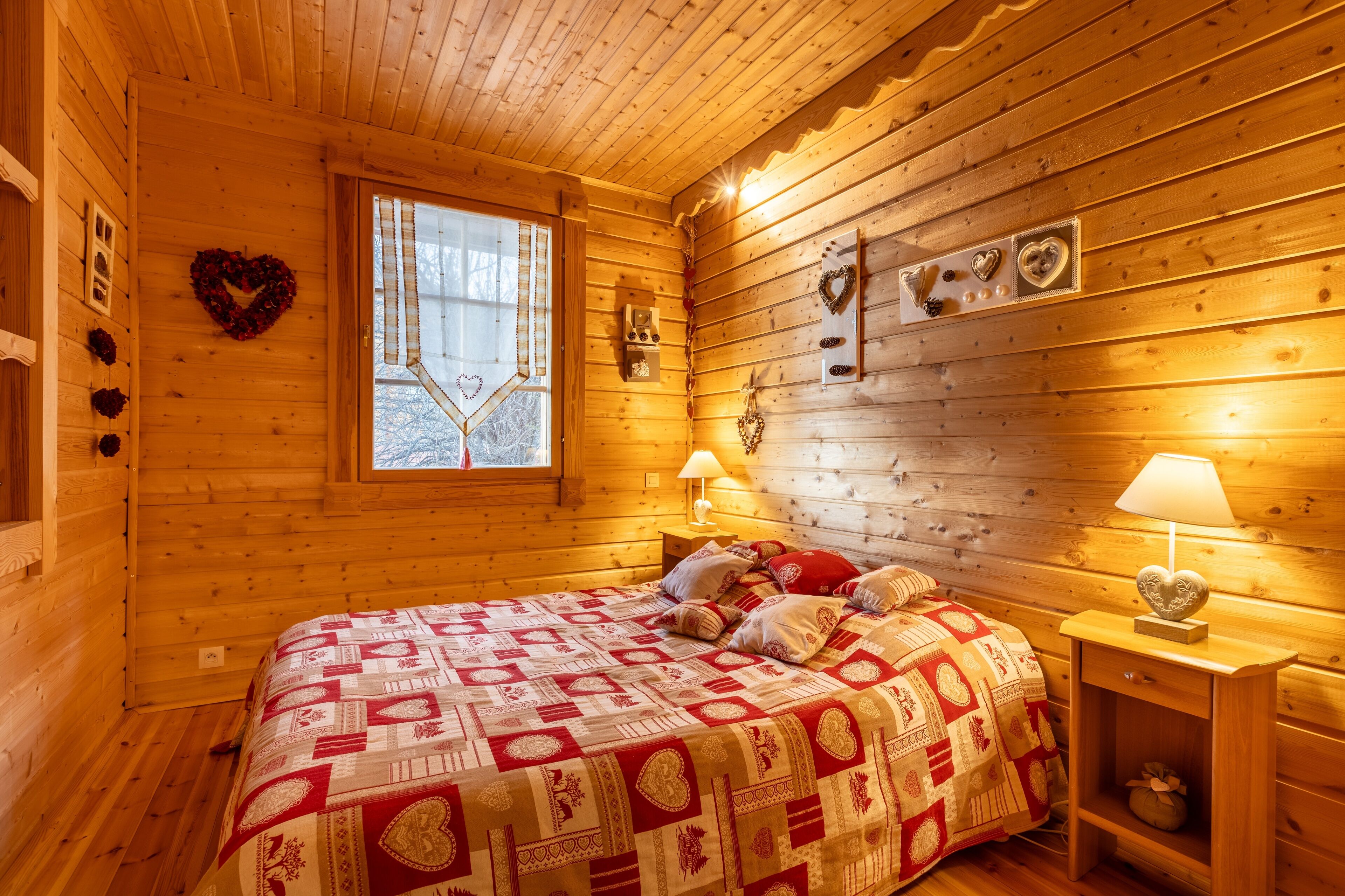 A cozy wooden cabin room with a bed, two bedside lamps, a window, and heart-shaped decorations on the walls.