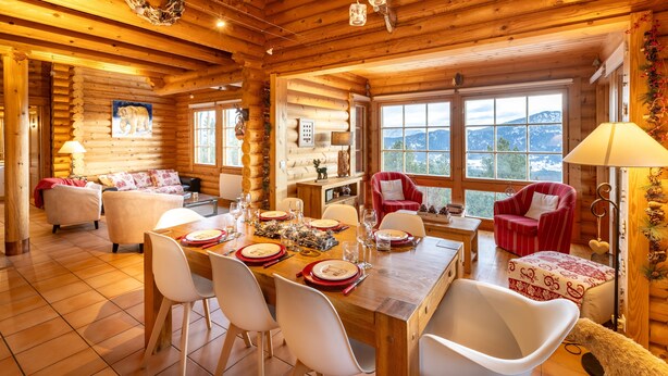 Interior de una cabaña de troncos con comedor, sala de estar con chimenea y vistas a montañas a través de amplias ventanas.