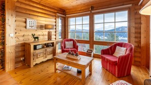 Interior acogedor de una cabaña de troncos con paredes de madera, un ventanal con vistas a la montaña, una mesa de centro de madera y dos sillones a rayas rojas.
