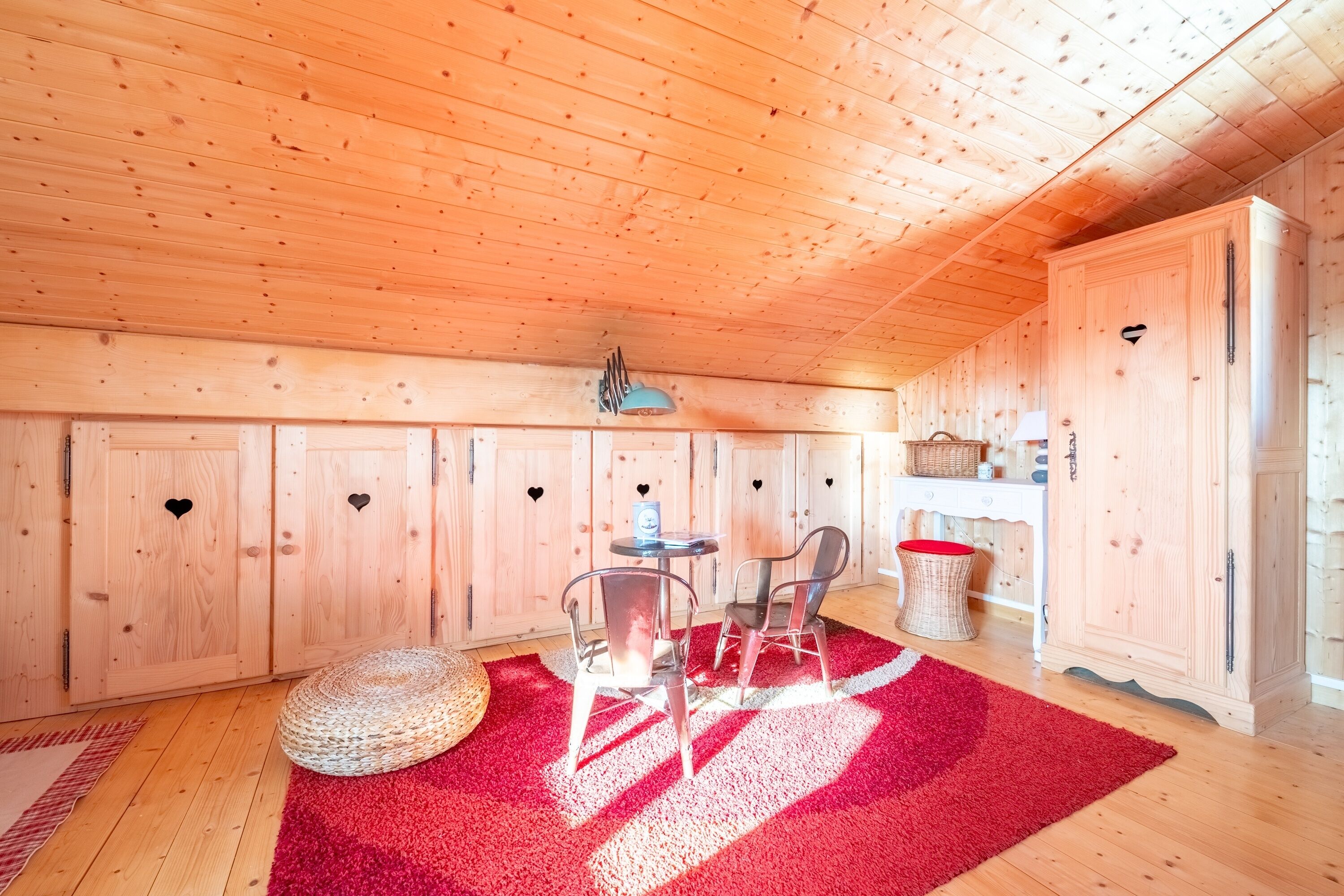 A wooden cabin interior with a red carpet, two chairs, a small table, and wooden cabinets with heart-shaped cutouts.