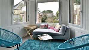 A cozy room with a grey sofa, two blue chairs, a small wooden table, and a window with a view of a stone building and trees.