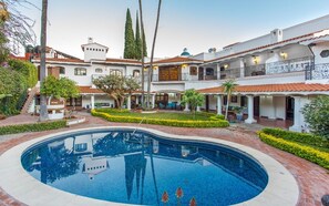 Uma piscina grande rodeada por um pátio com vários edifícios, incluindo uma estrutura de dois andares com varandas e um espaço coberto com terraço.