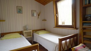 A room with two single beds, a small bedside table with a lamp, a window with a lace curtain, and a bookshelf with books. - Studio Avec Véranda Dans Vallée Calme à Reynès (Reynes)
