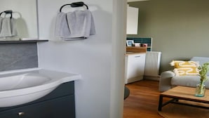 Um banheiro moderno com pia branca e armários escuros, uma toalha pendurada em um porta-toalhas, e um piso de madeira que leva a uma sala de estar com sofá e mesa de centro.