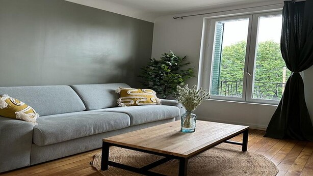 Uma sala de estar moderna com um sofá cinzento, uma mesa de centro de madeira e uma janela grande com cortinas.