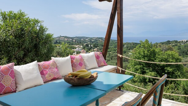 Une table bleue avec un saladier rempli de bananes et un banc en bois agrémenté de coussins à motifs.