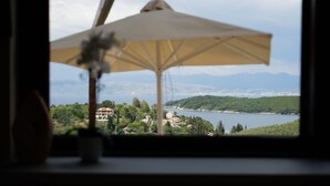 Une vue à travers une fenêtre, avec un grand parapluie, une plante en pot et un lac dont les montagnes se dessinent à l’arrière-plan.