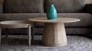 Une table ronde dotée d’un socle texturé et surmontée d’un vase, placée sur un tapis à motifs devant un canapé gris.