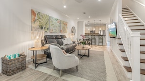 Ein geräumiges Wohnzimmer mit Sofa, Sessel und Couchtisch, darüber eine Treppe, im Hintergrund eine Küche.