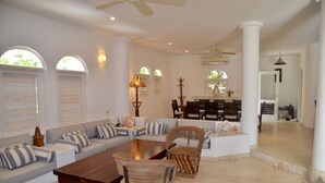 A spacious living room with a large sectional sofa, a wooden coffee table, and wicker chairs. There are two large windows with shutters, a ceiling fan, and a dining area in the background. - Christmas Deal!Oceanview private pool villa, steps from the beach. 24hs security (Playa del Carmen)