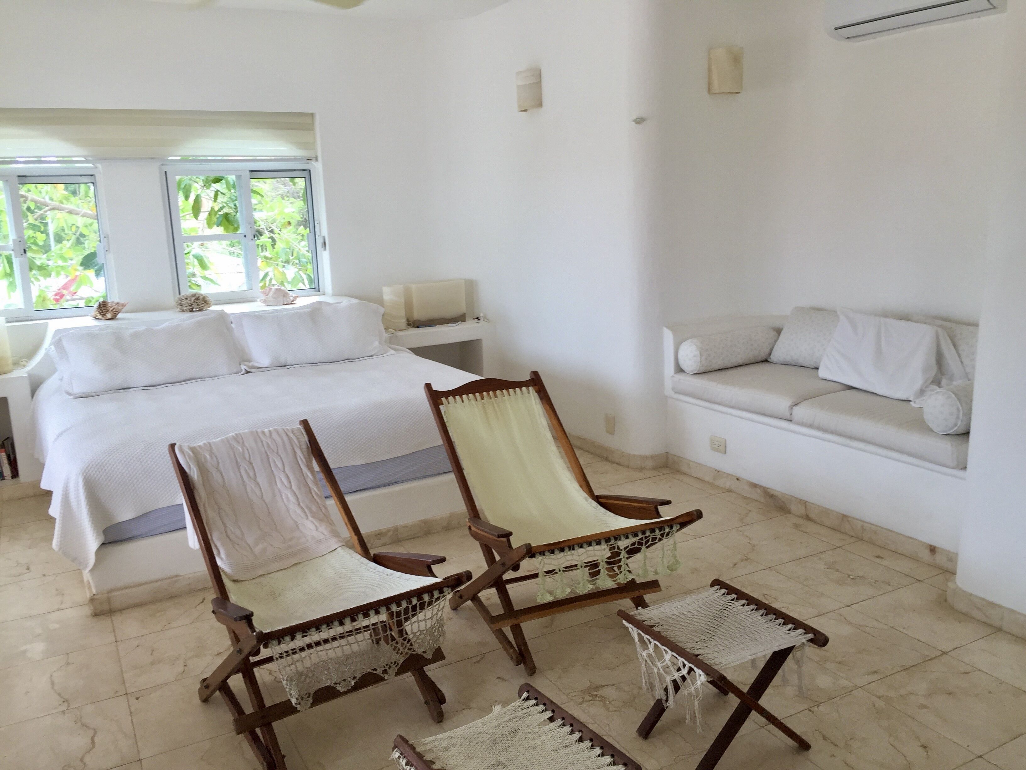 Un dormitorio con cama, dos sillas de madera con cojines blancos, un sofá blanco y una ventana con vistas al follaje.