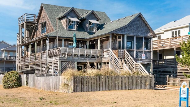 Une grande maison en bois à plusieurs étages, dotée d’un balcon et d’un porche couvert, et entourée d’une clôture en bois.