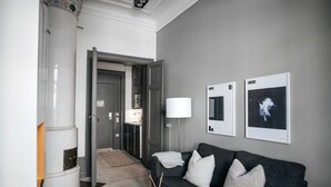 A modern living room with a dark grey sofa, a white side table, and a lamp. There are framed pictures on the wall and a door leading to another room. - Guldgränd  (Stockholm)