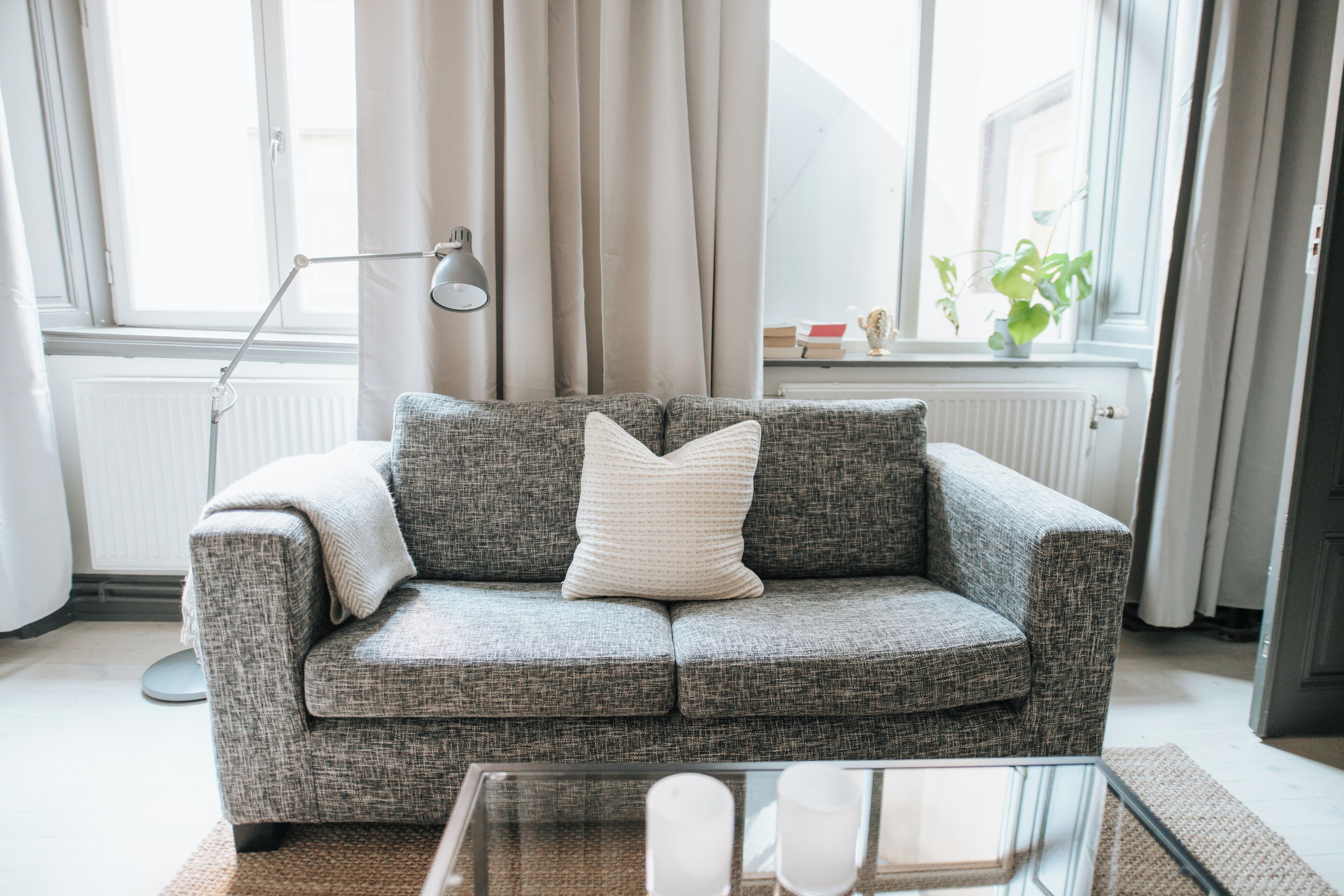 A grey sofa with a white pillow, a glass coffee table, a floor lamp, and a window with curtains.