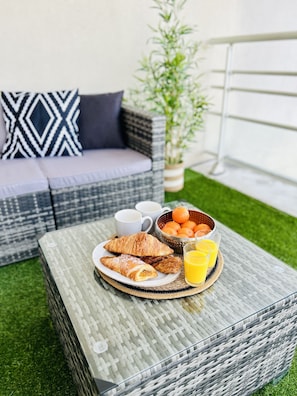Une table en verre est dressée pour le petit-déjeuner, avec des croissants, un saladier rempli d’oranges et des verres de jus d’orange.