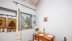A dining area with a wooden table, chairs, a window with curtains, and a view of trees outside. - Aimilia Cottage - One Bedroom Apartment, Sleeps 2 (Longos)