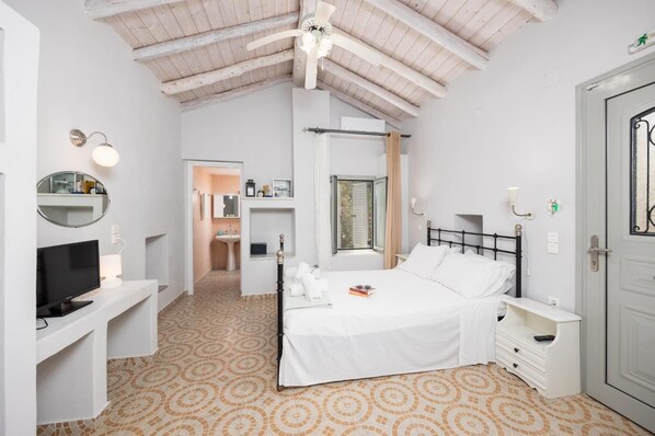 A bedroom with a bed, a TV, a desk, a mirror, a bathroom, and a window. - Aimilia Cottage - One Bedroom Apartment, Sleeps 2 (Longos)