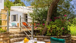 A dining area with a table set for two, a bottle of wine, and a bowl of lemons. There is a chair with a green cushion and a table with a floral tablecloth. - Aimilia Cottage - One Bedroom Apartment, Sleeps 2 (Longos)