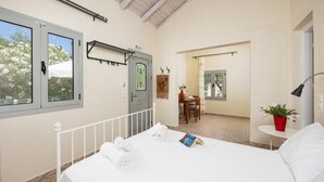A bedroom with a bed, a small table with a chair, a window with a view of trees, and a door with a towel rack. - Eleni Cottage - One Bedroom Apartment, Sleeps 2 (Paxi)