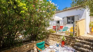 An outdoor dining area with a table set for two, surrounded by stone walls and a white building with shutters. - Eleni Cottage - One Bedroom Apartment, Sleeps 2 (Paxi)