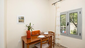 A small dining area with a wooden table, four chairs, a vase with flowers, and a window with curtains. - Eleni Cottage - One Bedroom Apartment, Sleeps 2 (Paxi)