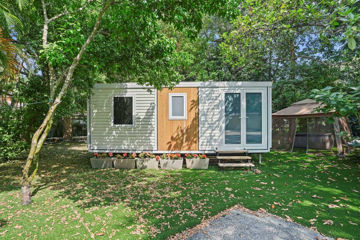 A small, single-story mobile home with a wooden door and windows, surrounded by a garden with potted plants and a lawn.
