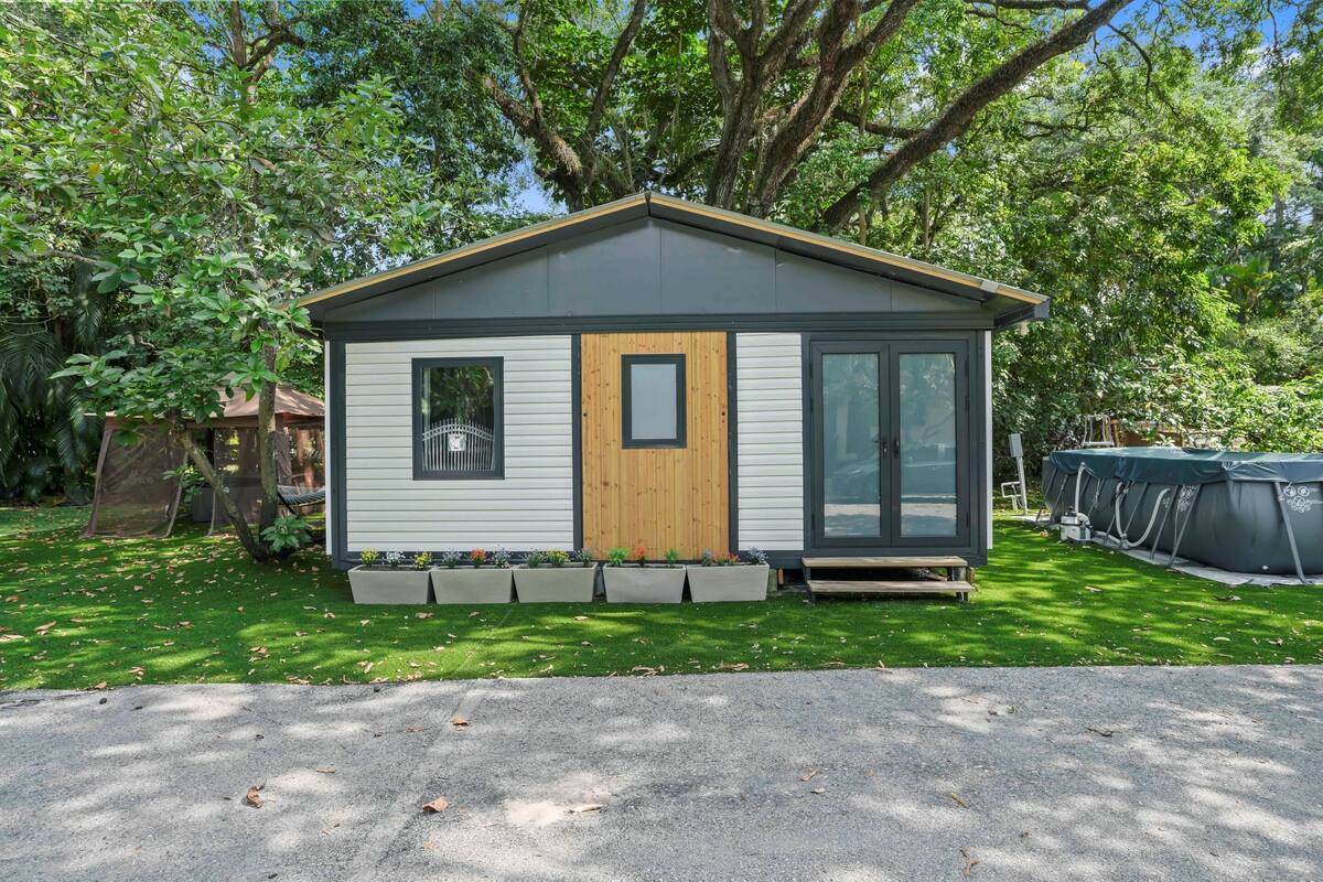 A small, single-story cabin with a wooden door and windows, surrounded by a well-maintained lawn and trees.