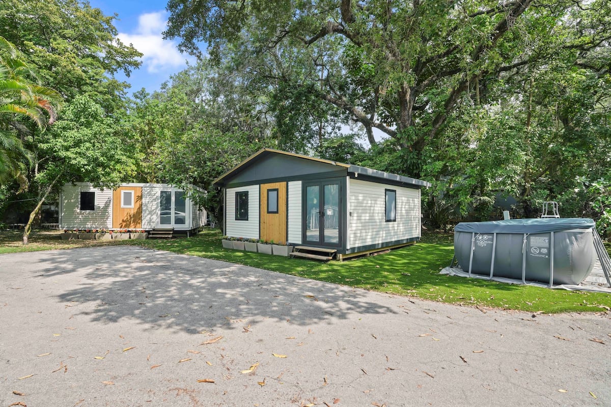 A small, single-story mobile home with a front porch and a sliding door, surrounded by lush greenery and a clear sky.